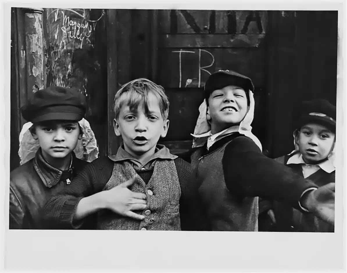 Helen Levitt Fotografía en blanco y negro de cuatro niños en una calle de Nueva York, tomada por la fotógrafa Helen Levitt en la década de 1940. Los niños, vestidos con ropa típica de la época, posan con expresiones vivas y gestos espontáneos frente a una puerta de madera, reflejando la energía y la inocencia de la infancia urbana en el Nueva York de mediados del siglo XX.