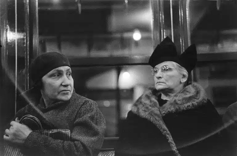 Walker Evans Fotografía en blanco y negro de dos mujeres sentadas en un vagón del metro de Nueva York. Ambas parecen ser mayores y están vestidas con abrigos formales. Una mujer lleva un sombrero oscuro y tiene una expresión pensativa mientras mira hacia el lado, mientras que la otra mujer, con gafas y un sombrero distintivo, mira hacia el frente con una expresión seria. La imagen captura una escena de la vida cotidiana con un estilo directo y analítico, típico de la fotografía de Evans.