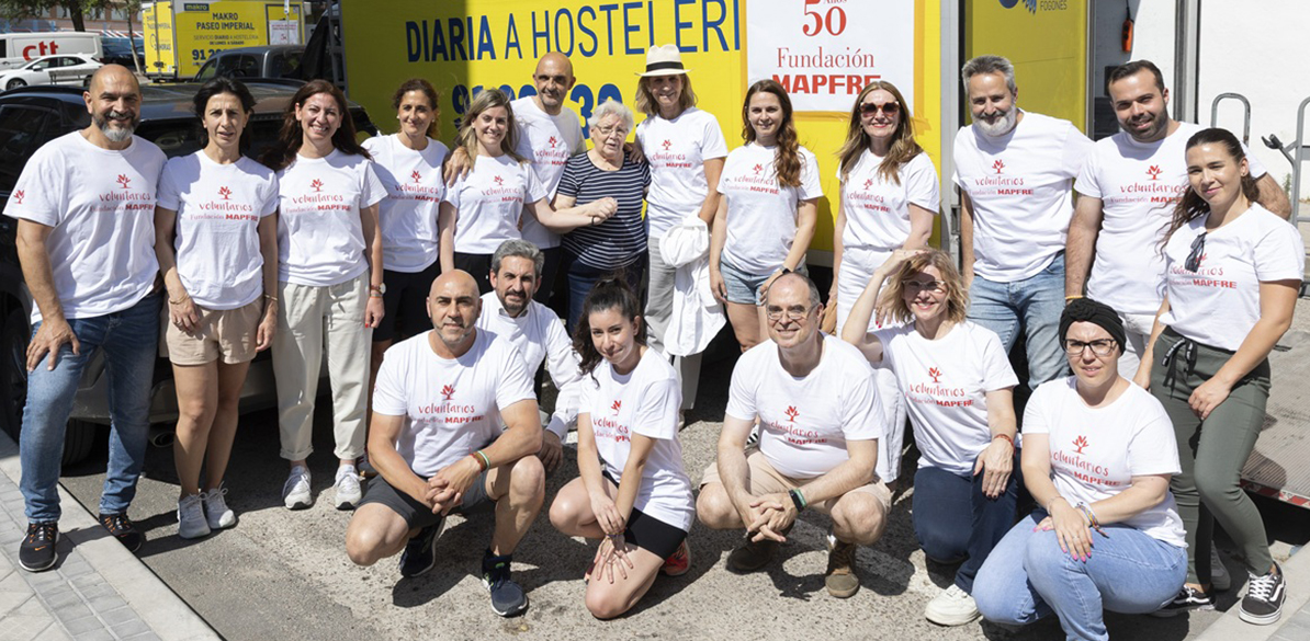 Pato Amarillo Grupo de 16 voluntarios de Fundación MAPFRE posando en una donación solidaria. todos llevan camisetas blanclas con el logo de "Voluntarios MAPFRE" en rojo, y están reunidos frente a una furgoneta amarilla.