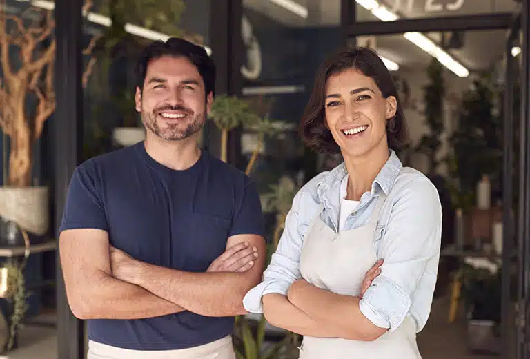 Grants for employment Two smiling shop owners stand confidently with arms crossed outside their plant store, surrounded by greenery. The image features earthy tones with beige, navy, and light blue clothing. Ideal for representing social action, collaboration, and sustainable small business values.